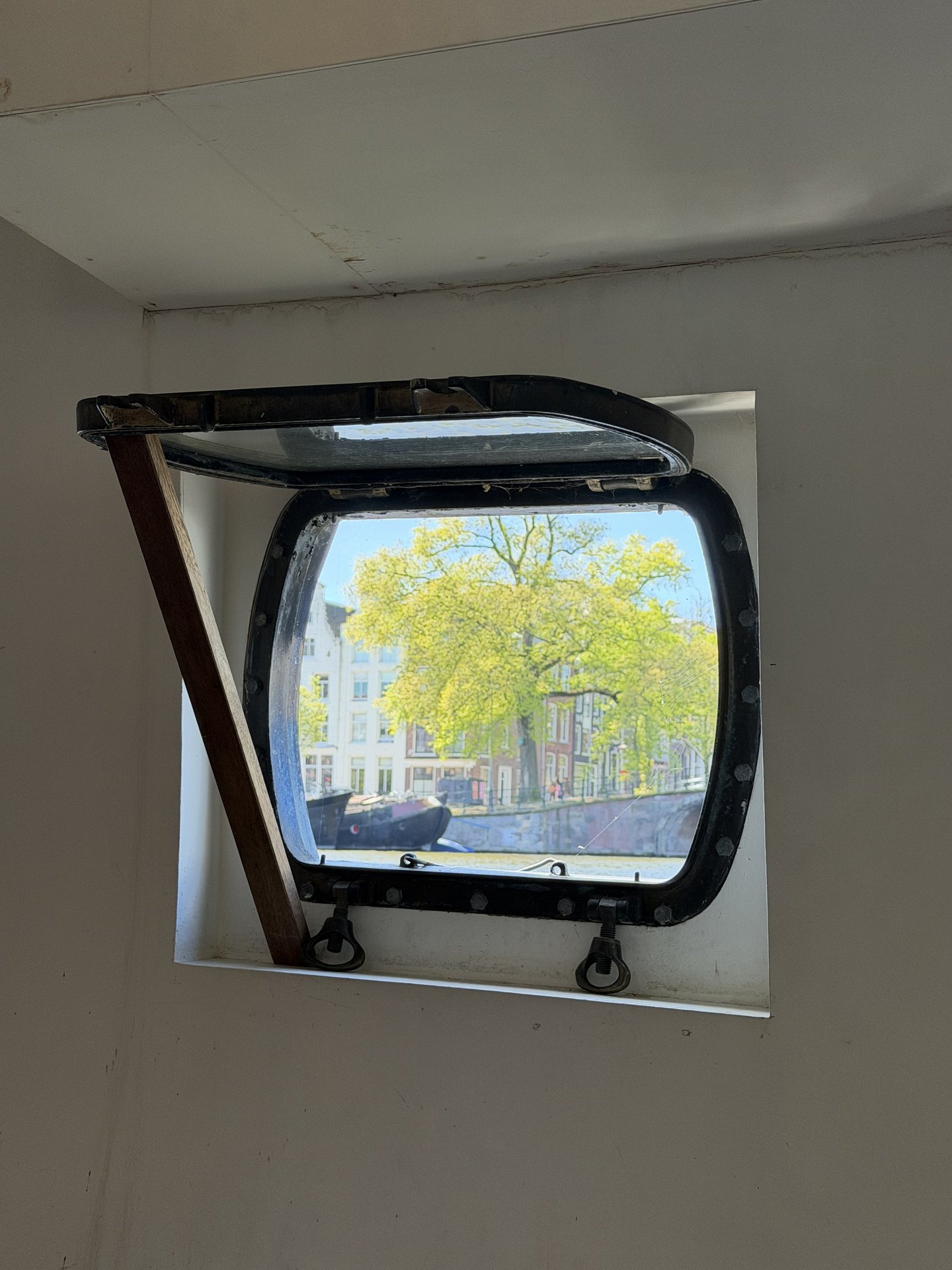 A unique porthole window, propped open with a wooden stick, offers a vibrant view of the Amstel canal in Weesperbuurt, Amsterdam